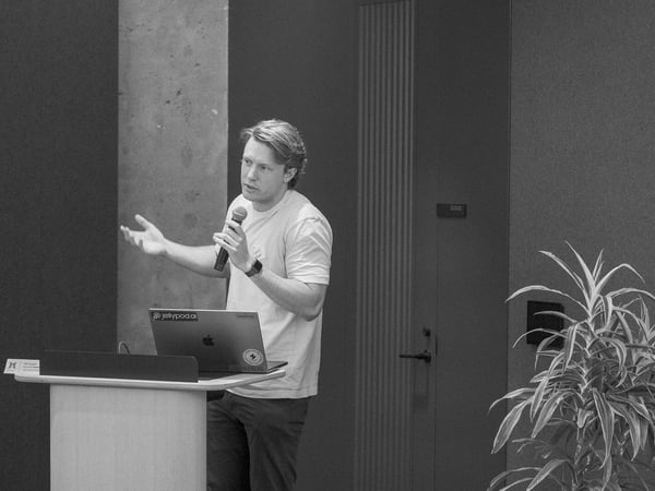 A black and white photograph of a man standing behind a podium, gesturing with his hand while speaking into a microphone during a presentation.