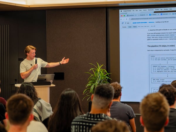 A man stands at a podium holding a microphone and gesturing towards a large projection screen displaying a GitHub repository page with a technical pipeline diagram to an audience.