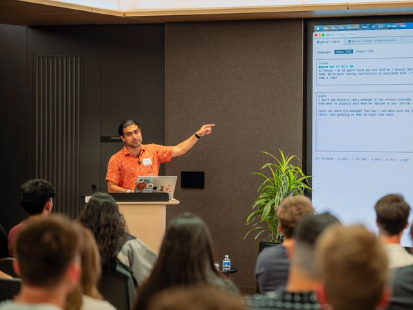 A man in an orange short-sleeved patterned shirt stands at a podium, pointing toward a large projection screen displaying an email or messaging application interface to an audience.