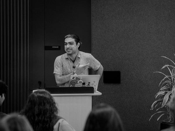 A black and white photograph of a man standing behind a podium with a sticker-covered laptop, giving a presentation to an audience.