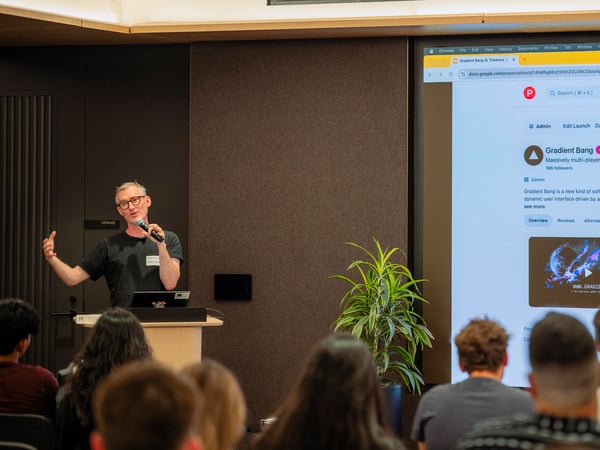 A man stands at a podium speaking into a microphone during a presentation, with a large screen behind him displaying a product page for 'Gradient Bang'.
