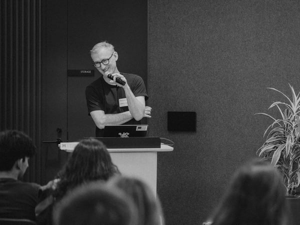 A black and white photograph of a man wearing glasses standing behind a podium, smiling and speaking into a microphone during a presentation.