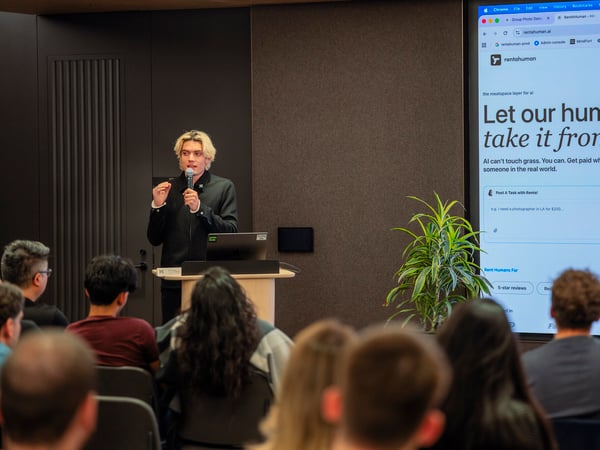 A young man with blonde hair stands at a podium giving a presentation, holding a microphone. Behind him, a large screen displays the website rentahuman.ai with the headline 'Let our humans take it from here', while an audience sits in the foreground watching him.