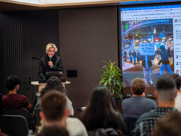 A young man with blonde hair speaks into a microphone at a podium during a presentation, with a large screen behind him displaying a social media post about an AI startup.