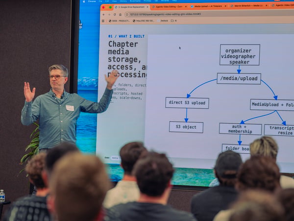 A man in a blue button-down shirt is giving a presentation in front of a large screen displaying a technical flowchart diagram about media storage and processing, while an audience sits in the foreground.