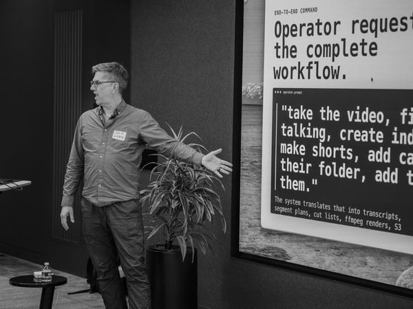 A black and white photograph of a man giving a presentation, gesturing towards a large screen displaying text about an operator request workflow.