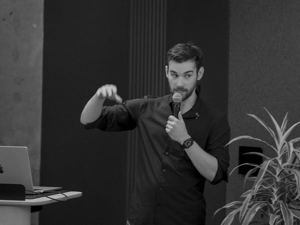 A black and white photograph of a man with a beard speaking into a microphone and gesturing with his hand during a presentation.