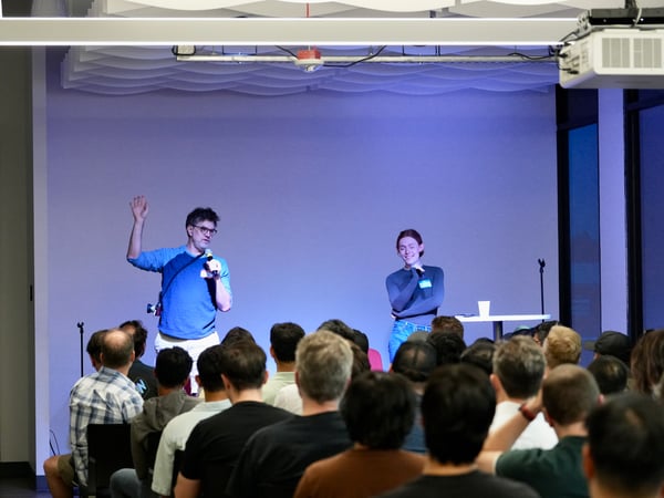 A photograph capturing an indoor event, likely a presentation or Q&A session, showing two presenters on a stage illuminated by blue light, addressing a large audience viewed from behind.