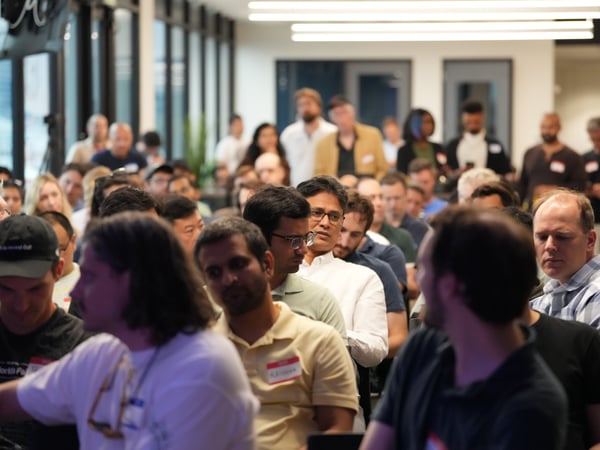 A candid photograph capturing a large audience attending an indoor event or presentation, with attendees seated closely together and focused toward the front of the room.