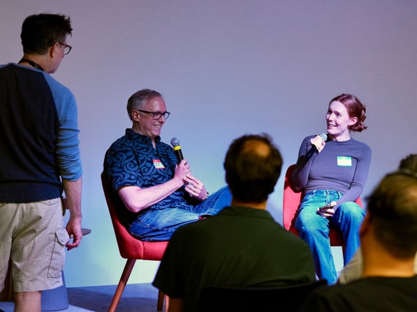 A candid photograph capturing a panel discussion at an indoor event, showing two seated speakers holding microphones and smiling, while several audience members watch from the foreground.