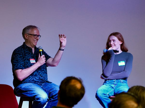 A man and a woman are seated on a stage, holding microphones, participating in a panel discussion or Q&A session, illuminated by dramatic purple stage lighting.