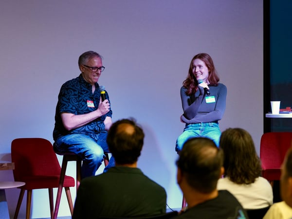 A photograph captures a panel discussion featuring a man and a woman seated on stools on a stage, both holding microphones and smiling at the audience. The background is a large, brightly lit screen or wall, and the foreground shows the backs of several audience members' heads.