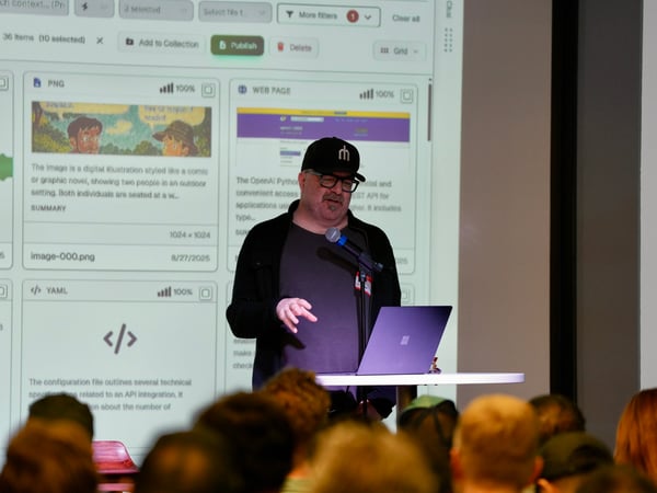 A man wearing a black hat and glasses is giving a technical presentation on stage, standing in front of a large projection screen displaying a content management interface with file previews and descriptions. The foreground shows the backs of several audience members' heads.