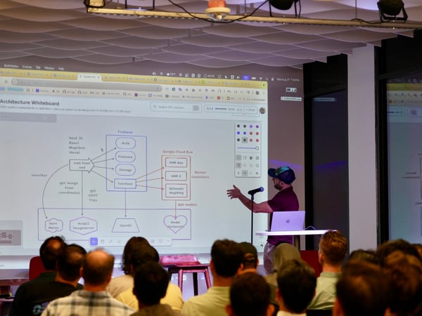 A photograph capturing an indoor technical presentation where a speaker, wearing a cap and a maroon shirt, gestures toward a projected screen displaying a detailed software architecture flowchart.