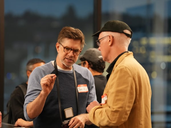 A candid photograph captures two men engaged in an animated conversation during an indoor evening event, with the man on the left wearing a name tag that reads 'Joe H.' and gesturing with his hand. The background features large windows overlooking a dark, blurry city skyline.