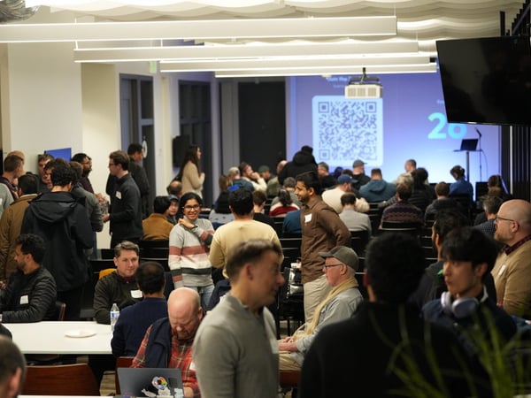 A candid photograph captures a crowded indoor professional or technical event, showing attendees mingling in the foreground and others seated facing a projection screen displaying a large QR code and the number 26 in the background.