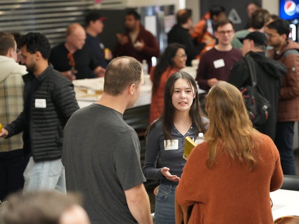 A candid photograph captures a crowded indoor networking or social event, focusing on a young woman speaking animatedly to two people whose backs are turned to the camera.