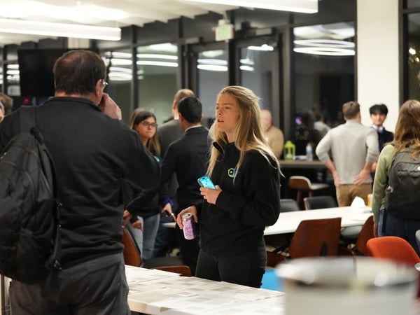 A candid photograph capturing a group of people mingling and networking indoors at a modern event space or office common area, featuring a woman in the foreground holding a phone and a beverage.