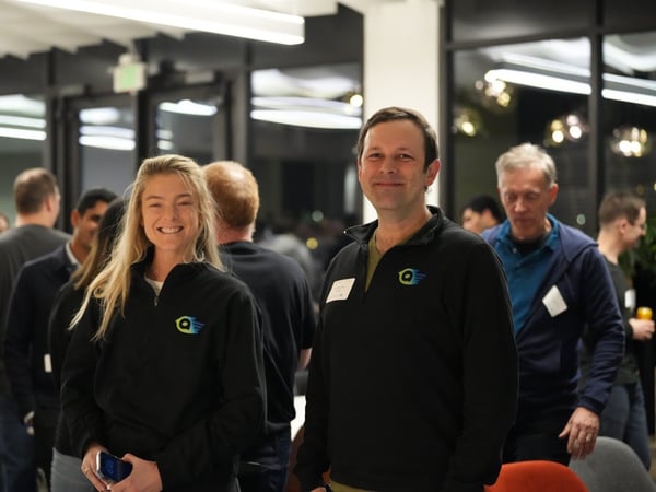 A candid indoor photograph capturing a smiling woman and man, both wearing black quarter-zip pullovers with a corporate logo, standing in the foreground of a crowded networking or social event.