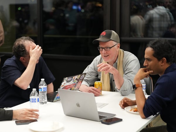 A candid photograph shows a group of men gathered around a white table indoors during an evening social or networking event, with open laptops, water bottles, and drinks visible.