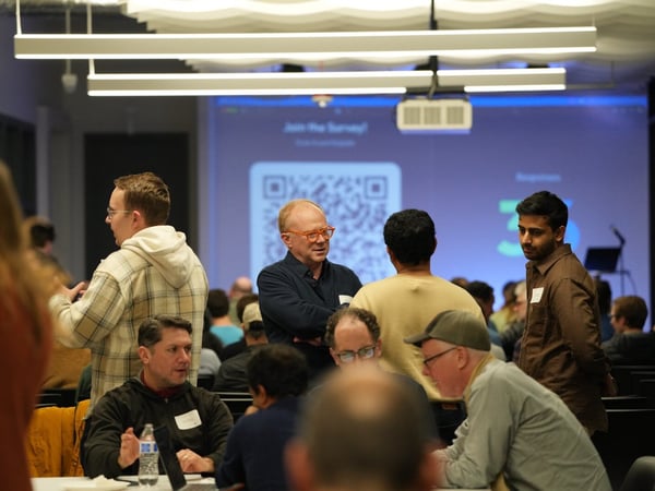 A candid photograph captures a group of attendees networking and conversing during an indoor event or conference, with a large screen displaying a QR code for a survey in the background.