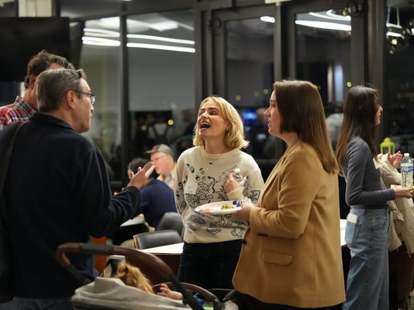 A candid photograph captures a group of people socializing and discussing at an indoor evening event, featuring a woman in a patterned sweater laughing or speaking animatedly in the center.