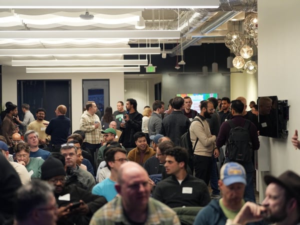 A candid photograph capturing a large indoor gathering, likely a tech meetup or presentation, showing dozens of attendees seated and standing in a modern office space with high ceilings and distinctive lighting fixtures.