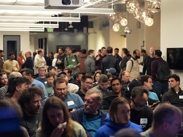 A large crowd of people, predominantly men, are gathered indoors, seated in the foreground and standing in the background, suggesting attendance at a professional meetup or presentation in a modern office space.