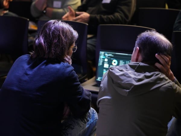 A candid photograph shows two individuals, viewed from behind, sitting closely together and intently focused on a glowing laptop screen in a dark, indoor conference or workshop setting.