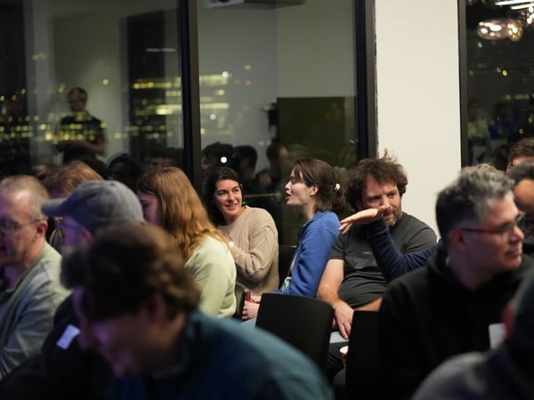 A low-light photograph captures a group of people seated in an indoor venue, likely attending a presentation or meeting, with a blurred cityscape visible through large windows in the background.