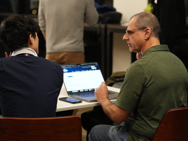A candid indoor photograph capturing two men seated at a table, engaged in a discussion while looking at the screen of an open laptop. A third person is visible standing in the blurred background.