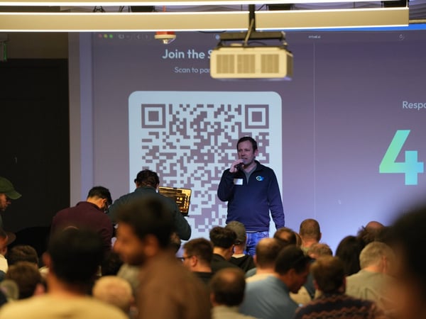 A man wearing a blue pullover speaks into a microphone while standing in front of a large screen displaying a QR code, presentation text, and the number '44' to a large, seated audience in an indoor setting.