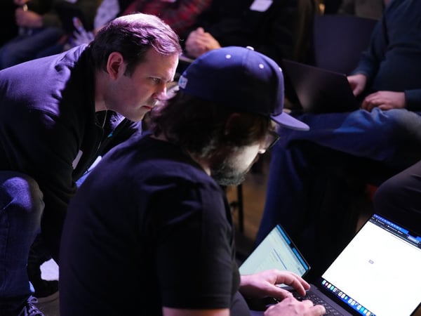 A candid photograph taken in a dark, purple-lit indoor setting, showing two men leaning over a laptop, seemingly collaborating or debugging code during a technical event or conference. Several other attendees are partially visible in the dimly lit background.
