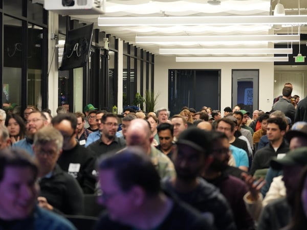 A candid photograph captures a large, densely packed audience attending an indoor presentation or event in a modern office space, with many attendees visible in the mid-ground and the foreground heavily blurred.