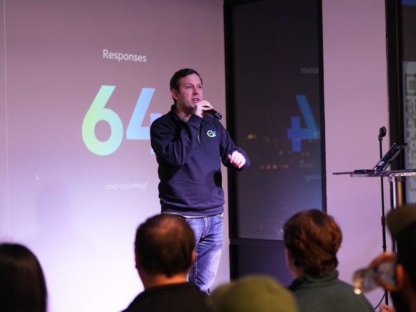 A man is giving a presentation on stage at an indoor event, holding a microphone in front of a large projection screen that displays the text 'Responses 64 and counting!' The backs of several audience members are visible in the foreground.