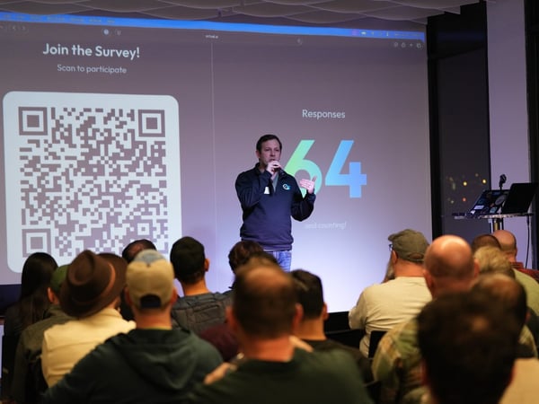 A photograph capturing a presentation event where a male speaker is addressing an audience in a dimly lit room, standing in front of a large screen displaying a QR code for a survey and a live response count of 64.