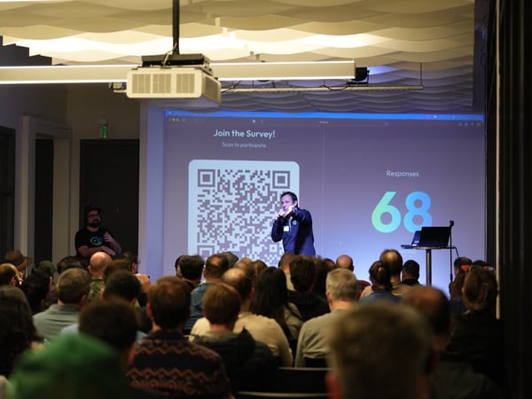 A photograph taken inside a modern venue shows a speaker presenting in front of a large screen displaying a QR code survey and the number 68 responses, while a large audience watches.