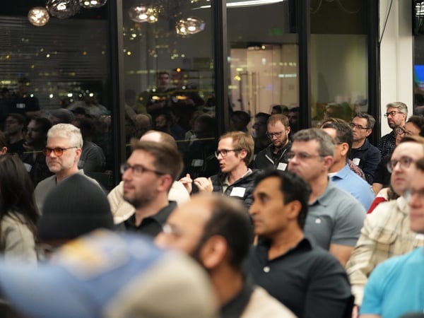 A candid photograph captures a large audience seated indoors during a nighttime event, with attendees looking toward a speaker or presentation off-camera, and city lights reflecting in the large windows behind them.