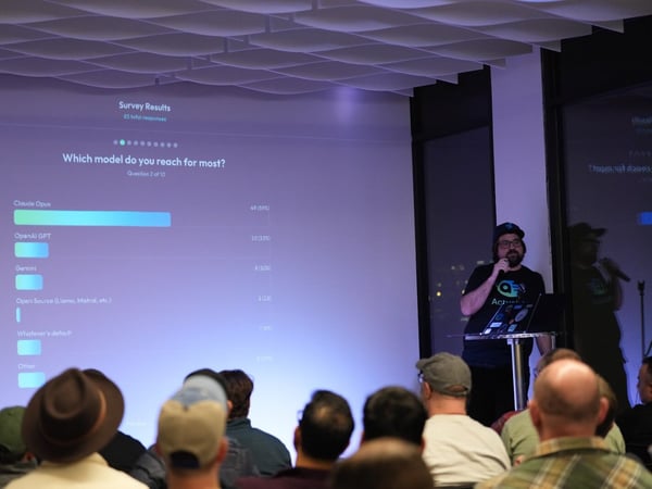 A photograph taken during an evening presentation where a speaker, dressed in a dark shirt and cap, stands beside a large screen displaying survey results about preferred AI models, while an audience watches from the foreground.