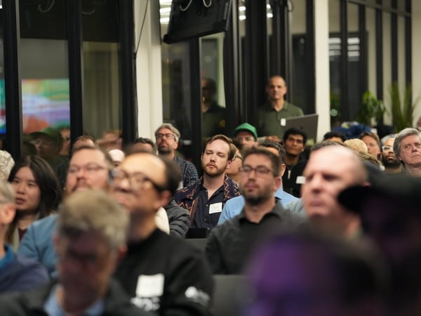 A candid photograph captures a large audience seated indoors during an event, with most attendees looking attentively towards an unseen speaker or screen off-camera. The image uses a shallow depth of field, rendering the immediate foreground heavily blurred while focusing on the middle rows of people.