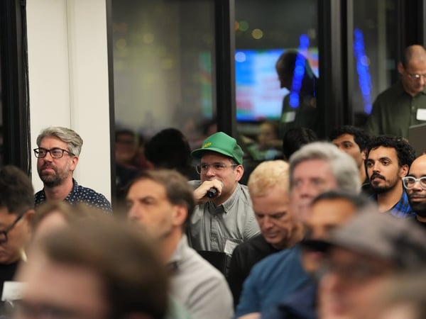 A candid photograph captures a seated audience attending an indoor event, likely a presentation or discussion, with several men in the mid-ground looking forward. One man wearing a green baseball cap is visible holding a microphone, suggesting he is speaking or asking a question.