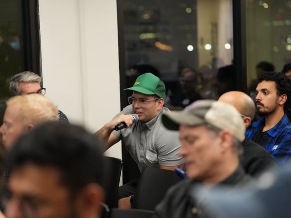 A man wearing a green baseball cap and glasses is speaking into a handheld microphone while seated in an audience during a dimly lit indoor event or Q&A session.