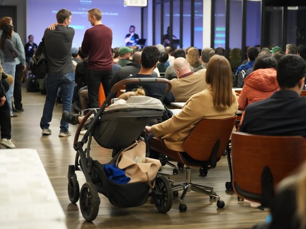 A candid photograph capturing a large audience seated indoors, attending a presentation or conference, with a baby stroller visible in the foreground.