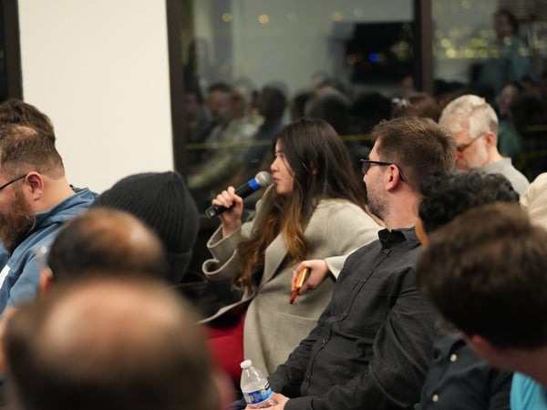 A candid photograph captures an indoor evening event showing an audience, with a woman in the center holding a microphone, suggesting she is asking a question or speaking during a Q&A session. The background features a large window reflecting the room and showing blurred city lights outside.