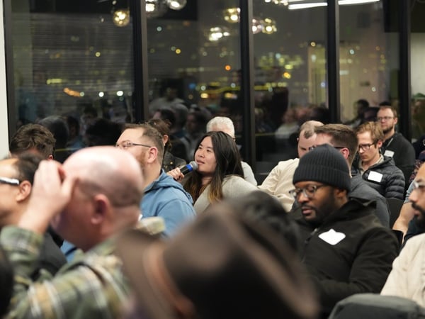 A candid photograph captures a diverse audience attending an indoor event at night, focusing on a woman in the center holding a microphone, suggesting a Q&A session or discussion. The background features large windows overlooking a blurred, illuminated city skyline.