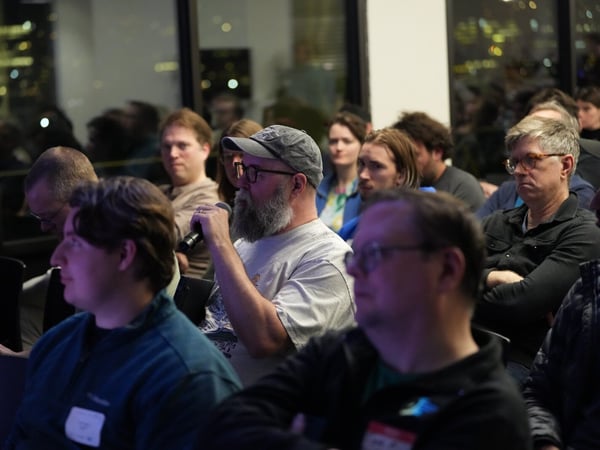 A candid photograph taken during a nighttime indoor event, showing an audience member, a bearded man wearing a cap, holding a microphone to speak or ask a question.