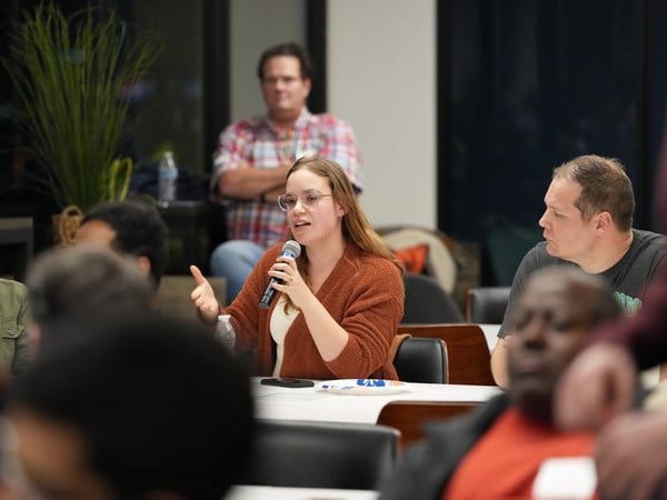 A young woman wearing glasses and a rust-colored cardigan speaks into a handheld microphone while seated at a table during an indoor group discussion or event.
