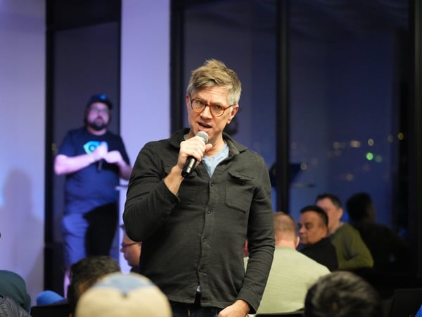 A man wearing glasses and a dark shirt speaks into a microphone during a nighttime indoor event, with a blurry audience in the foreground and city lights visible through large windows in the background.