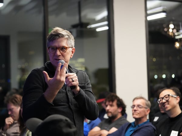 A middle-aged man wearing glasses and a dark shirt is speaking into a microphone and gesturing with his hand during an indoor presentation or Q&A session, with several audience members visible in the dimly lit background.