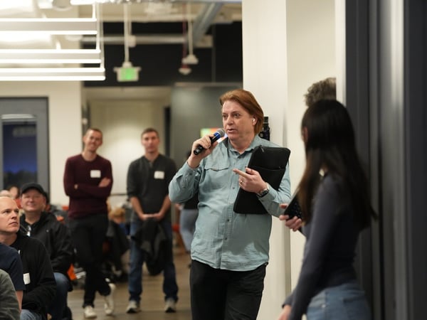 A man with reddish hair is speaking into a microphone while holding a black folder, participating in a discussion or Q&A session during an indoor professional event.